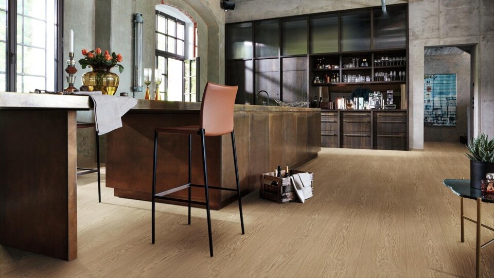 Laminatboden Edition m8 mit Endlosoptik in heller Eiche-Optik unter einer dunkelbraunen Kücheninsel mit Barhocker